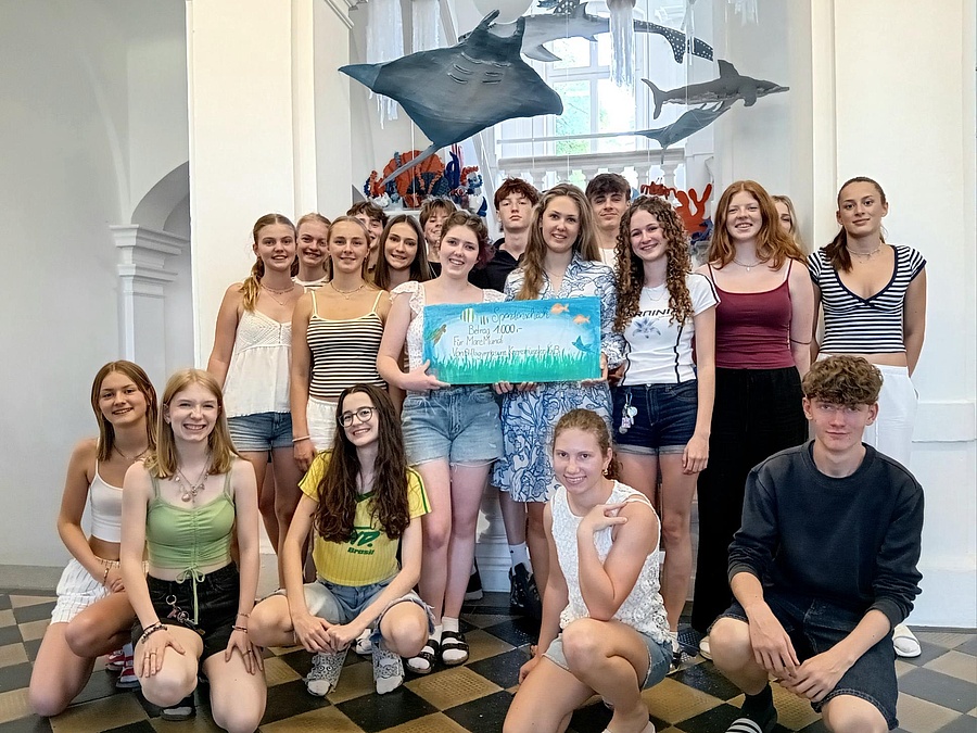 Die Schüler:innen der 6B mit dem Spendenscheck vor der Unterwasserwelt im Stiegenhaus des Stiftsgymnasiums Kremsmünster.