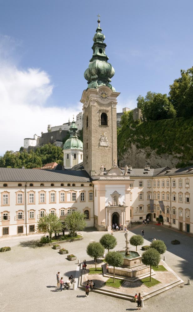Blick in den äußeren Stiftshof mit Kirchenfassade, Turm und Vierungskuppel sowie Brunnen; im Hintergrund die Festung Hohensalzburg