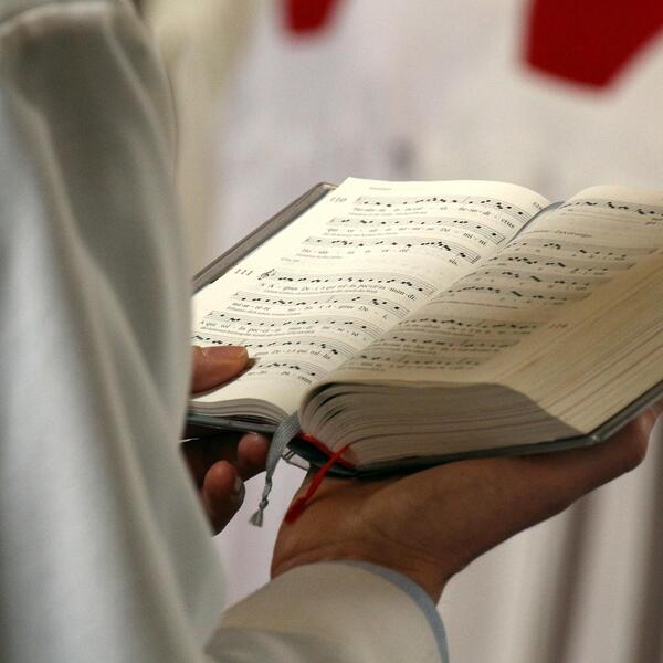 Offene Hand hält ein Gesangbuch, im Hintergrund Ministranten